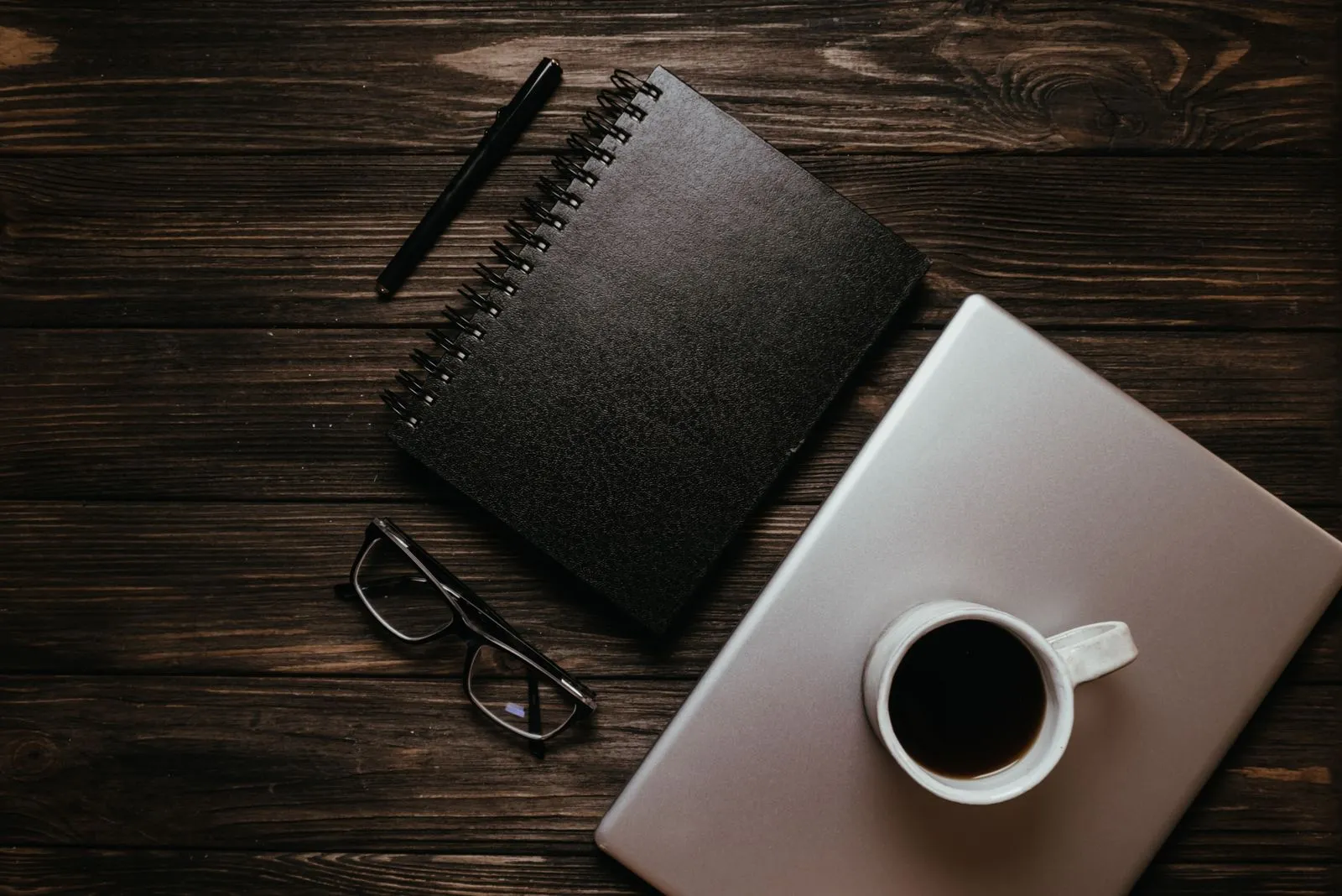 Black notebook and pen beside a laptop and coffee on a dark wooden table