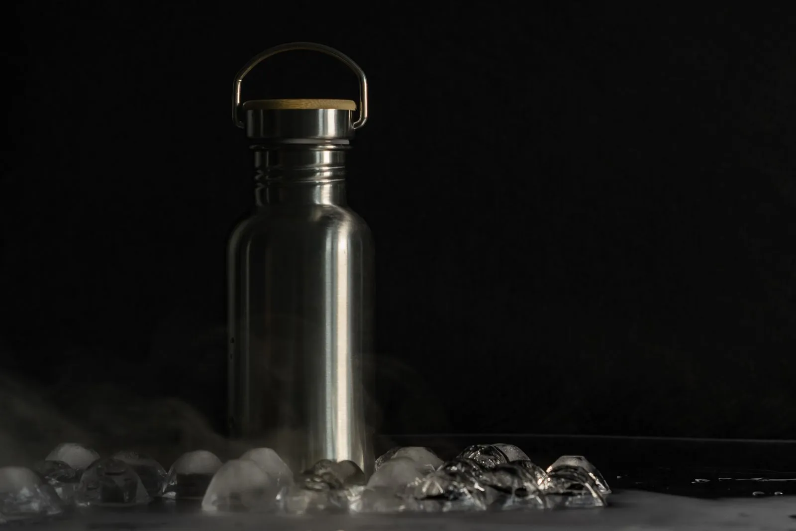 Stainless steel water bottle with a bamboo lid among ice on a black background